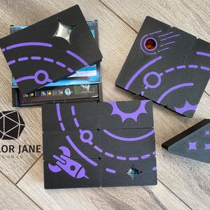 May include: A set of black foam tiles with purple space-themed designs. The tiles are arranged in a grid pattern and feature images of planets, stars, rockets, and comets. The tiles are designed to be used as a game board or for decorative purposes.