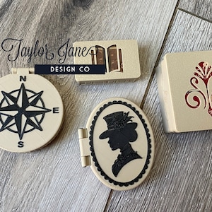May include: Three small, beige, decorative boxes with different designs. The first box has a compass design with a black compass rose. The second box has a silhouette of a woman wearing a hat. The third box has a red, ornate design.