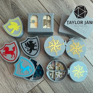 May include: A collection of gray, shield-shaped game pieces with various animal emblems, along with compass-themed containers and a castle-shaped box. The pieces are in shades of gray, yellow, red, and blue. The text "TAYLOR JANE DESIGN CO." is visible.