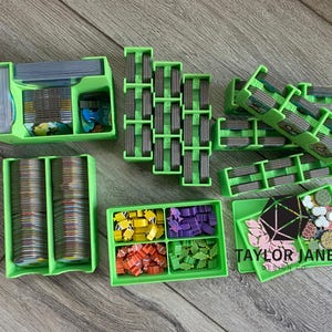 May include: A collection of bright green game organizers, including card holders, token trays, and component organizers. The organizers are filled with game pieces, cards, and tokens in various colors. The text "TAYLOR JANE" is visible on one of the trays.