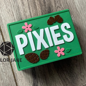May include: A green rectangular sign with the word "PIXIES" in large white letters. The sign is decorated with pink flowers, brown acorns, and leaves. The Taylor Jane Design logo is in the lower left corner.