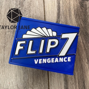 May include: A blue rectangular object with the text "FLIP 7 VENGEANCE" in white. The object has a white fan-like design above the word "FLIP". The logo "TAYLOR JANE DESIGN CO" is in the upper left corner.