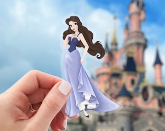 May include: A sticker featuring a cartoon illustration of a woman with long brown hair wearing a blue and purple dress. The sticker is held in a hand in front of a blurred background of a pink castle.
