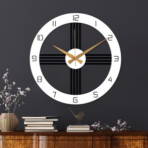 May include: A circular wall clock featuring a white outer ring with black Arabic numerals from 1 to 12. The centre has a white circular hub with gold-coloured hands and black horizontal and vertical lines extending to the outer ring.