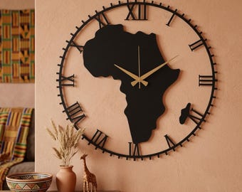 May include: A large circular wall clock featuring a black metal frame with Roman numerals and a central silhouette of the African continent. The clock has thin, gold-colored metal hands that contrast against the black map shape.
