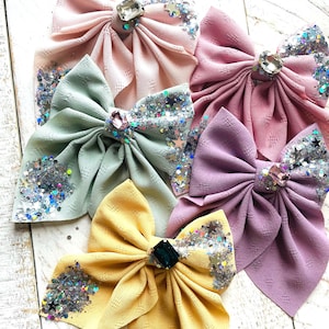 May include: Five fabric hair bows with glitter and jeweled accents. The bows are in shades of pink, green, purple, and yellow.