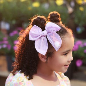 May include: A young girl with curly brown hair wearing a light pink and white floral dress with a lavender bow in her hair. The bow has a pink bunny applique with a white fluffy tail.