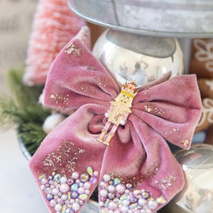 May include: A pink velvet bow with a gold nutcracker ornament in the center. The bow is decorated with iridescent beads and glitter.