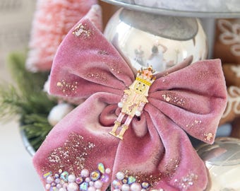 May include: A pink velvet bow with a gold nutcracker ornament in the center. The bow is decorated with iridescent beads and glitter.