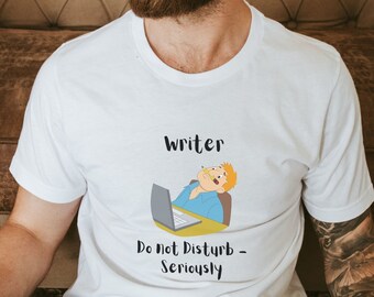 May include: White t-shirt with a cartoon illustration of a person working on a laptop. The text "Writer Do Not Disturb - Seriously" is printed on the shirt.