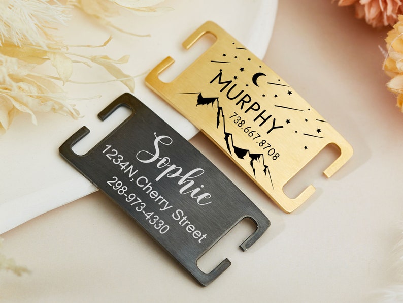 May include: Two personalized pet ID tags. One is gold with a mountain and stars design and the text "MURPHY 738.667.8708". The other is black with the text "Sophie 1234N, Cherry Street 298-973-4330".