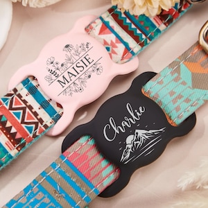 May include: Two colorful dog collars with personalized name tags. The pink tag has a floral design and the text "Maisie". The black tag has a mountain design and the text "Charlie".