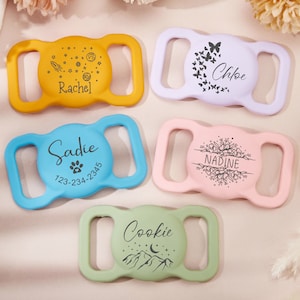 May include: Five colorful plastic dog collar buckles with personalized engravings. The buckles feature different designs, including a rocket ship, butterflies, a paw print, flowers, and a mountain range. The names "Rachel", "Chloe", "Sadie", "Nadine", and "Cookie" are engraved on the buckles.