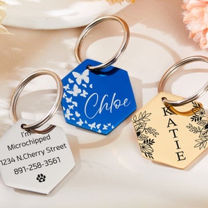 Puede incluir: Dos etiquetas de identificación para mascotas personalizadas. Una es plateada con texto negro que dice "I'm Microchipped 1234 N. Cherry Street 891-258-3561" y una huella de pata. La otra es azul con mariposas blancas y el nombre "Chloe" en letra blanca.