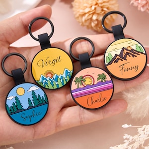 Pode incluir: Quatro chaveiros redondos com alças e argolas pretas. Cada chaveiro apresenta um design de paisagem diferente com um nome. Os designs incluem montanhas, árvores e um pôr do sol. Os nomes são Virgil, Fanny, Charlie e Sophie.