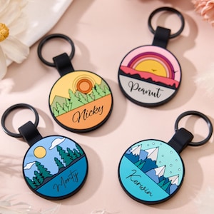 Pode incluir: Quatro chaveiros redondos com correias e argolas pretas. Cada chaveiro apresenta um design de paisagem diferente com um nome. Os designs incluem um pôr do sol, um arco-íris, montanhas e árvores. Os nomes são Nicky, Peanut, Monty e Kerwin.