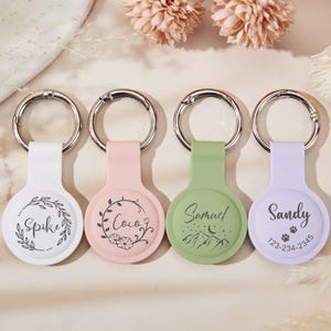 May include: Four AirTag holders in white, pink, green, and lavender, each with a silver keyring. The white holder has the name "Spike" with a leaf design. The pink holder has the name "Coco" with a floral design. The green holder has the name "Samuel" with a mountain design. The lavender holder has the name "Sandy" with a paw print and phone number.