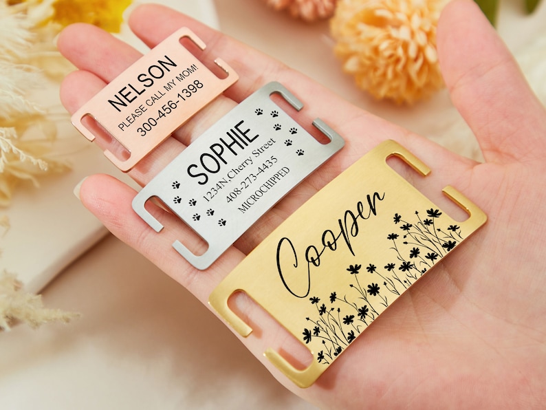 May include: Three personalized dog tags with different designs. The first tag is rose gold and says "NELSON PLEASE CALL MY MOM! 300-456-1398". The second tag is silver and says "SOPHIE 1234N, Cherry Street 408-273-4435 MICROCHIPPED" with paw prints. The third tag is gold and says "Cooper" with a floral design.
