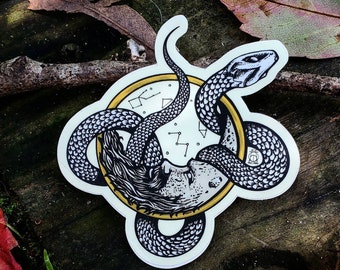 Op de afbeelding: Een zwart-witte sticker van een slang die om een ​​klokgezicht met een gouden achtergrond is gewikkeld. Het klokgezicht heeft een sterrenbeeldontwerp.