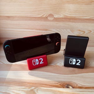 Könnte beinhalten: Eine schwarze Handheld-Spielkonsole mit roten und blauen Akzenten, die auf einem roten Ständer mit dem Text "2" ruht. Ein schwarzer Ständer mit dem Text "2" ist ebenfalls vorhanden. Die Artikel befinden sich auf einer hellen Holzoberfläche.