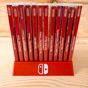 Puede incluir: Un soporte rojo para juegos de Nintendo Switch con varias carcasas de juegos. Las carcasas rojas con texto blanco muestran títulos como "Mario Kart 8 Deluxe" y "Super Mario Odyssey". El soporte tiene el logotipo de Nintendo Switch.