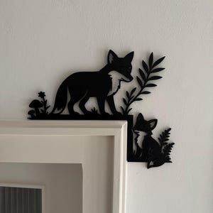 Op de afbeelding: Zwarte metalen vos wanddecoratie. De set bevat een groot vos silhouet dat op een grasachtige basis met een paddenstoel staat en een kleinere vos die met bladeren zit. De decoratie is gemonteerd op een witte muur en een wit deurkozijn.