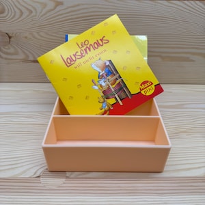 May include: A peach-colored rectangular organizer with two compartments holds a children's book titled "Leo Lausemaus will nicht essen." The book has a yellow cover with illustrations of a mouse and other characters. The background is a light wood grain.