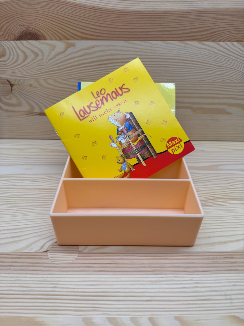 May include: A peach-colored rectangular organizer with two compartments holds a children's book titled "Leo Lausemaus will nicht essen." The book has a yellow cover with illustrations of a mouse and other characters. The background is a light wood grain.