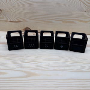 May include: Five small, black plastic battery organizers, each with a handle and labeled with battery types: AA, AAA, 9V, and two unlabeled. The organizers are arranged on a light wood surface.