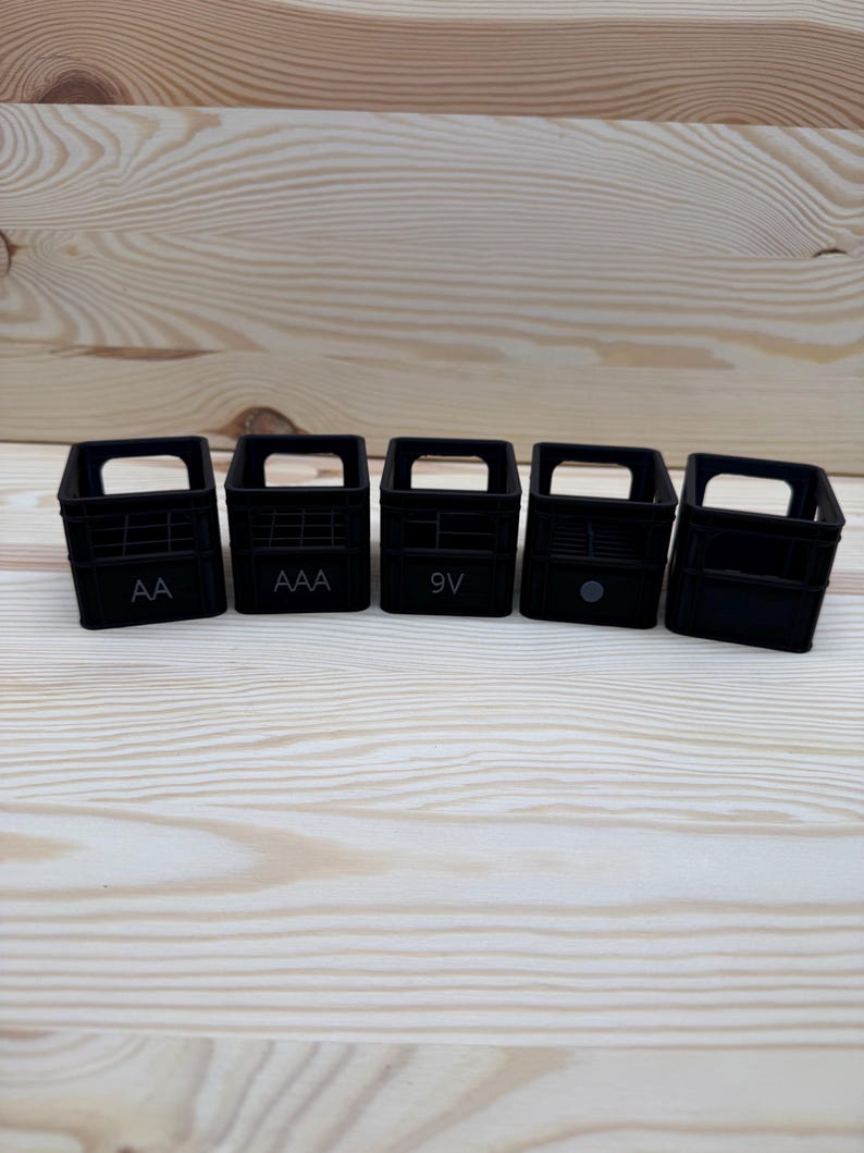 May include: Five small, black plastic battery organizers, each with a handle and labeled with battery types: AA, AAA, 9V, and two unlabeled. The organizers are arranged on a light wood surface.