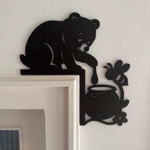 Op de afbeelding: Zwart berensilhouet dat naar honing reikt die uit een pot druppelt, met een bij en bloem accent. De beer zit op een richel en de honingpot staat in een hoek. Decoratieve wandkunst.