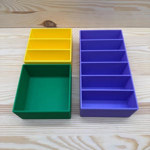 May include: Four colorful plastic organizers in yellow, green, and purple. The yellow organizer has three compartments, the purple has five, and the green is a square tray. These are ideal for desk organization.