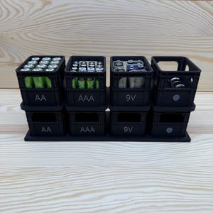 May include: Black battery organizer with four stacked compartments. Each compartment is labeled with battery type: AA, AAA, 9V, and a fourth for miscellaneous batteries. The organizer is designed to hold and display batteries.