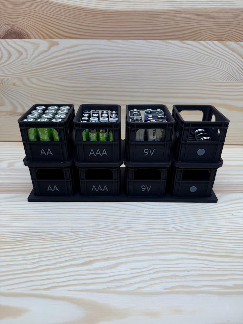 May include: Black battery organizer with four stacked compartments. Each compartment is labeled with battery type: AA, AAA, 9V, and a fourth for miscellaneous batteries. The organizer is designed to hold and display batteries.