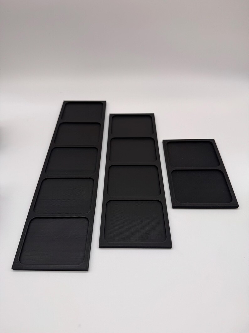 May include: Three black rectangular trays with multiple compartments. The trays are arranged in a row, varying in size and number of compartments. The trays are made of a matte black material and are set against a white background.