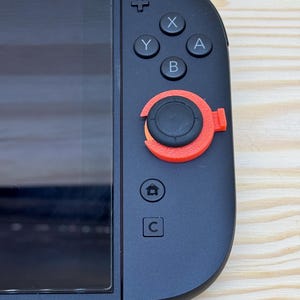 Op de afbeelding: Een zwarte gamecontroller met een scherm aan de linkerkant. De controller heeft een rood-zwarte joystick en de knoppen X, Y, A, B, een home-knop en een C-knop. De controller ligt op een licht houten oppervlak.