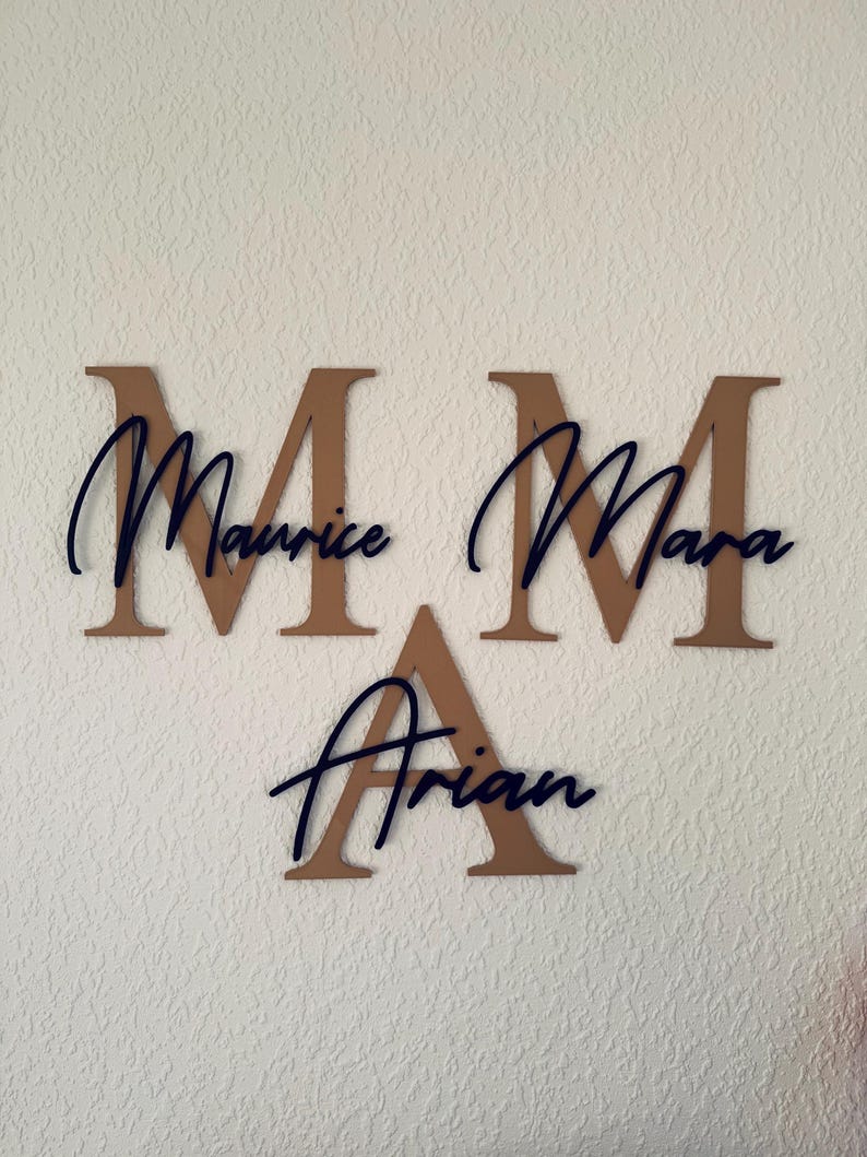 Op de afbeelding: Drie houten muurbrieven die de namen Maurice, Mara en Arian spellen. De grote, bruine hoofdletters zijn gecombineerd met de namen in een donkerblauwe, cursieve letter. De letters zijn gemonteerd op een getextureerde, gebroken witte muur.