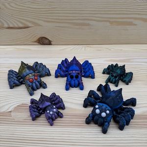 Può includere: Cinque figurine di ragni colorati, in tonalità di blu, viola e verde, sono disposte su una superficie di legno chiaro. Ogni ragno ha un design unico, con zampe e occhi dettagliati. I ragni variano per dimensioni e colore.