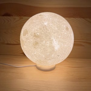 Op de afbeelding: Een bolvormige lamp met maantextuur, die een zacht, warm licht uitstraalt. De lamp staat op een kleine, cirkelvormige basis, met een dunne witte kabel. De achtergrond is van hout.