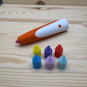 Könnte beinhalten: Ein orange-weißer 3D-Stift mit rosa Spitze, zusammen mit sechs farbigen Stiftdüsen in Blau, Gelb, Lila, Rot und Hellblau. Die Artikel sind auf einer hellen Holzoberfläche angeordnet.