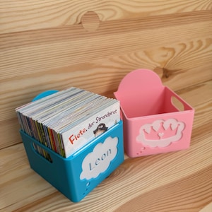 Può includere: Due contenitori in plastica, uno blu e uno rosa, su una superficie di legno. Il contenitore blu contiene libri e il nome "Leon". Il contenitore rosa presenta un disegno a corona.