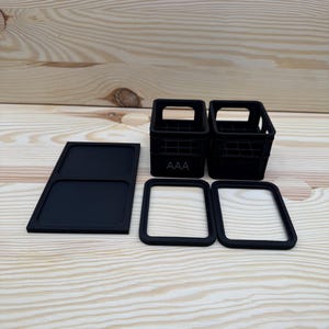 May include: Black miniature crates, a tray, and two square frames are arranged on a light wood surface. The crates have a grid design and the letters "AAA" on the front. The frames and tray are solid black.