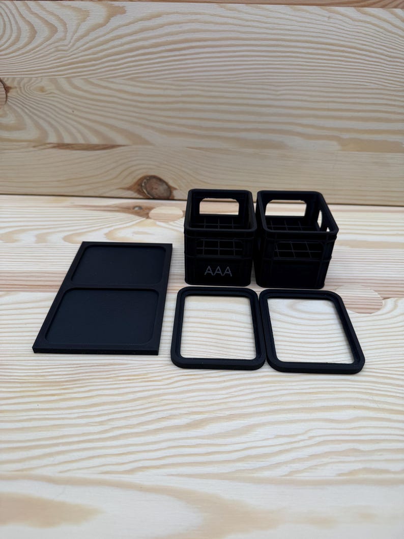 May include: Black miniature crates, a tray, and two square frames are arranged on a light wood surface. The crates have a grid design and the letters "AAA" on the front. The frames and tray are solid black.