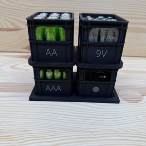 May include: A black 3D-printed battery organizer with compartments for AA, AAA, and 9V batteries. The organizer is stacked and sits on a base. The AA compartment has "AA" printed on it, and the 9V compartment has "9V" printed on it.