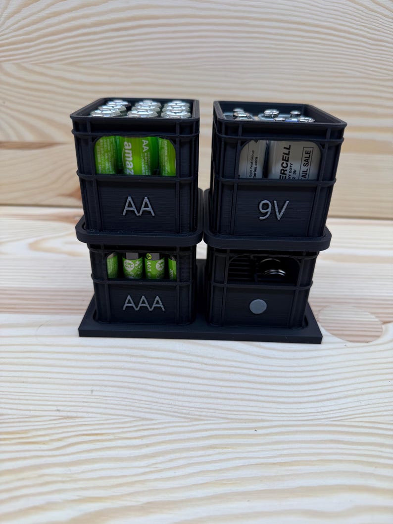 May include: A black 3D-printed battery organizer with compartments for AA, AAA, and 9V batteries. The organizer is stacked and sits on a base. The AA compartment has "AA" printed on it, and the 9V compartment has "9V" printed on it.