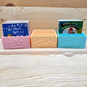 Può includere: Tre piccoli contenitori in plastica colorati, ciascuno contenente una pila di libri per bambini. I contenitori sono rosa pastello, pesca e blu, con decorazioni floreali, solari e lunari. I libri hanno testo tedesco sulle loro copertine.