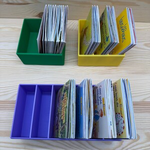 May include: Three colorful book organizers filled with children's books. A green organizer holds a stack of books, a yellow organizer holds a few books, and a purple organizer has multiple compartments with books.