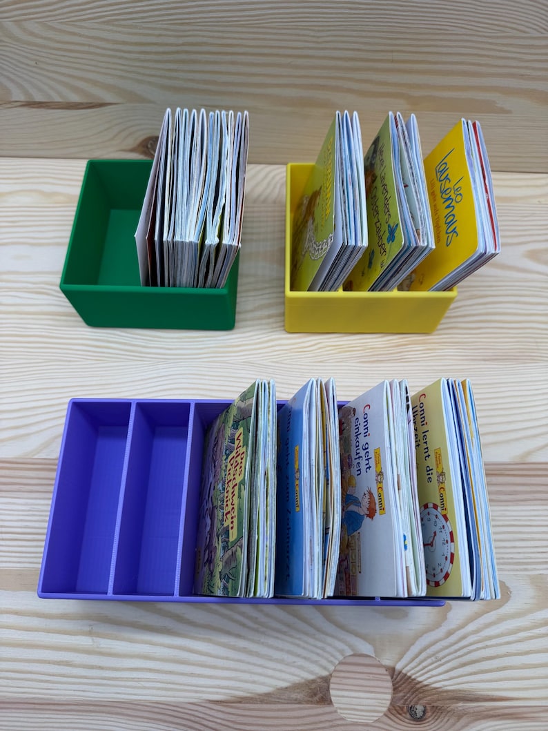 May include: Three colorful book organizers filled with children's books. A green organizer holds a stack of books, a yellow organizer holds a few books, and a purple organizer has multiple compartments with books.