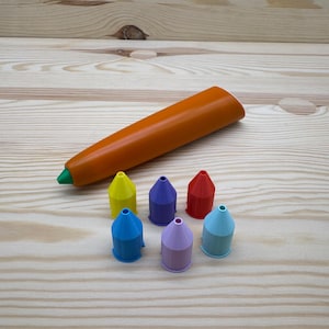Könnte beinhalten: Ein orangefarbener Stift mit grüner Spitze liegt neben sechs farbigen, kegelförmigen Objekten. Die Objekte sind in Blau-, Gelb-, Lila- und Rottönen gehalten. Die Artikel sind auf einer hellen Holzoberfläche angeordnet.