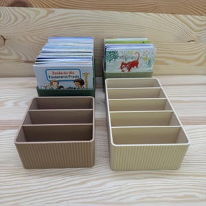 Può includere: Quattro organizer in plastica beige e verde con lati a coste verticali. Due organizer sono riempiti con pile di piccole carte. Le carte nell'organizer verde hanno il testo "Entdecke die Kinderarzt-Praxis".